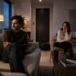 Two adults sit far apart in a living room as one prepares to leave, conveying emotional distance at the end of a relationship.