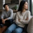 Two adults sit apart in a living room, one withdrawn and the other looking concerned.