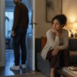 A couple sits apart in a modern apartment, with one person standing near an open doorway while the other looks uncertain on a sofa.