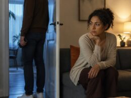 A couple sits apart in a modern apartment, with one person standing near an open doorway while the other looks uncertain on a sofa.